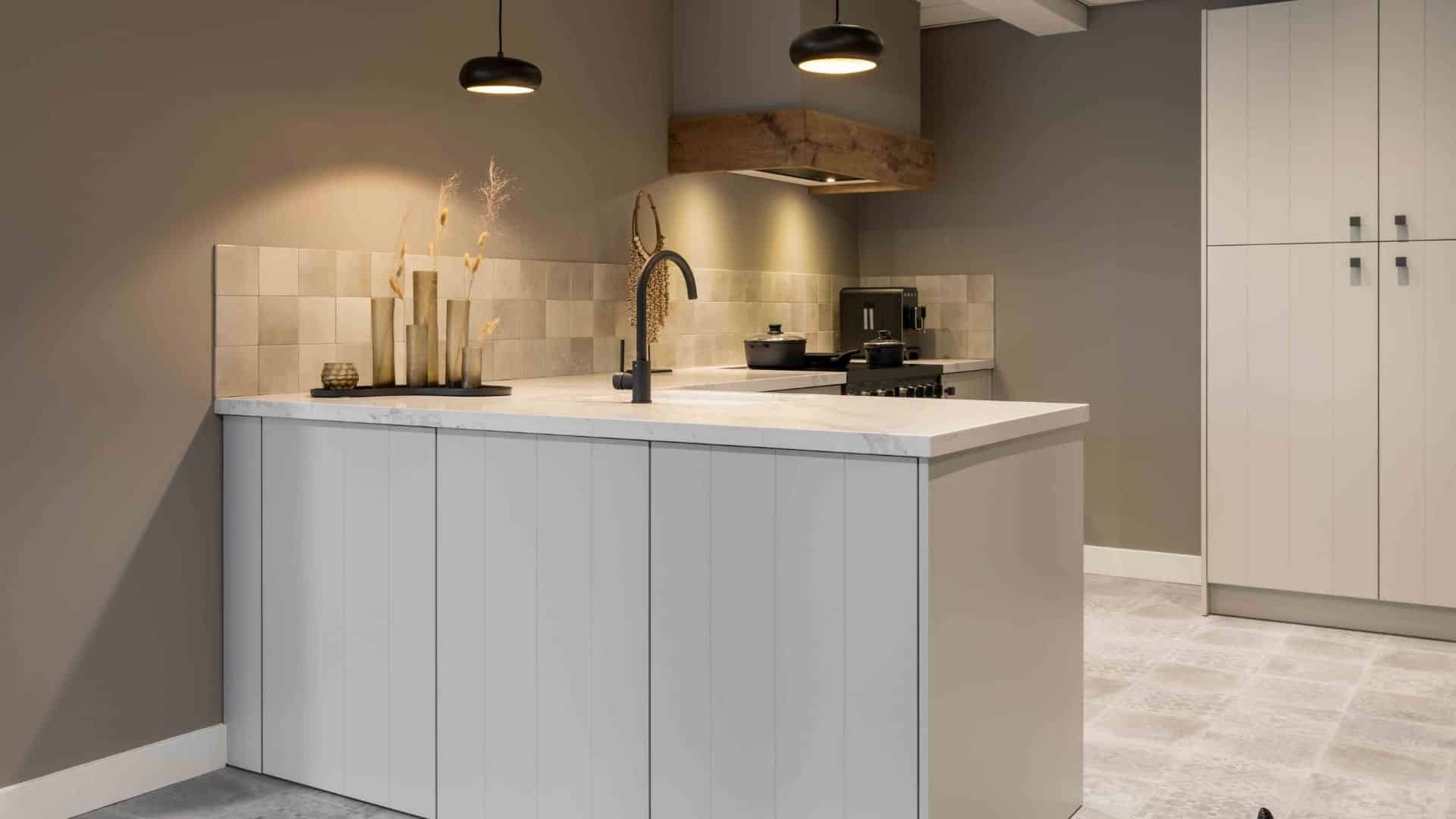 Impressie van een keuken in hoekopstelling. De keuken heeft geen bovenkasten, maar een eiland met lades en een kast op keuken spullen op te bergen. Het eiland wordt verlicht door twee design plafondlampen.