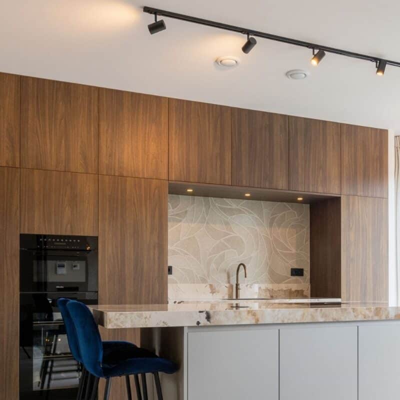 Impressie van een keuken met noten houten bovenkastjes. Onder de bovenkastjes zien we een nis en inbouw keuken apparatuur.