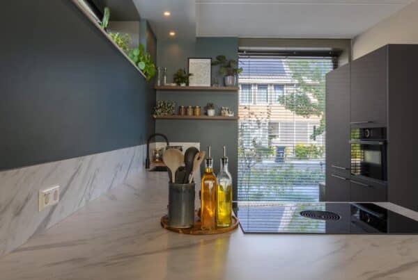 Impressie van een moderne hoekkeuken met natuursteenlook blad, kookplaat met werkbladafzuiging en zwarte kastenwand met inbouw apparatuur.