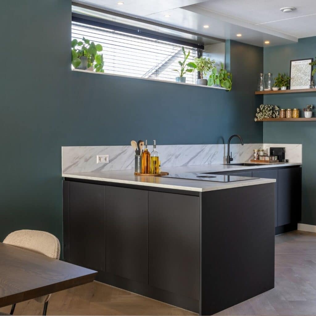 Impressie van een moderne keuken met hoekopstelling. De keuken is voorzien van zwarte kasten en een natuursteenlook werkblad in wit. Het blad loopt een stukje door op de groene wand.