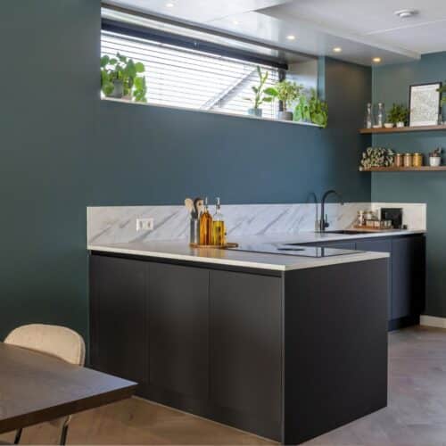 Impressie van een moderne keuken met hoekopstelling. De keuken is voorzien van zwarte kasten en een natuursteenlook werkblad in wit. Het blad loopt een stukje door op de groene wand.