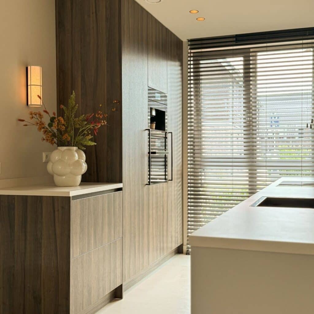 Impressie van een Japandi keuken met een houten kastenwand, voorzien van inbouw keukenapparatuur. Een wandlamp voor warme sfeerverlichting.