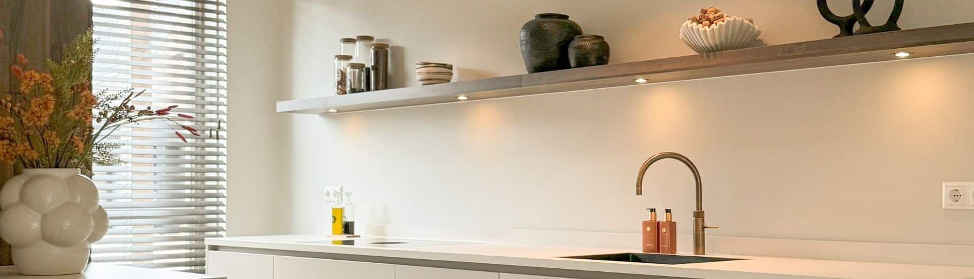 Impressie van een Japandi keuken in een parallel opstelling. De keuken is ingericht met een strak werkblad, veel opberglades, een Quooker, een wijnklimaatkast en houten kastenwand.