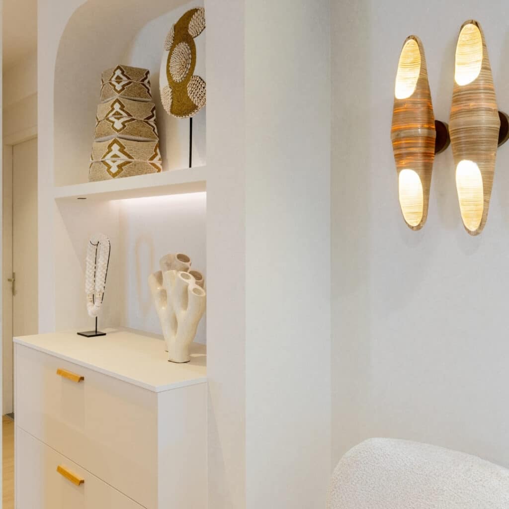 Impressie van een deel van een witte Japandi keuken. We zien een nis met ronde hoeken, met de uitstraling van een toog. De nis is voorzien van twee onderkasten met houten handgrepen, met daarboven een verlichte plank met decoraties. Naast de nis zien we twee organisch gevormde wandlampen.