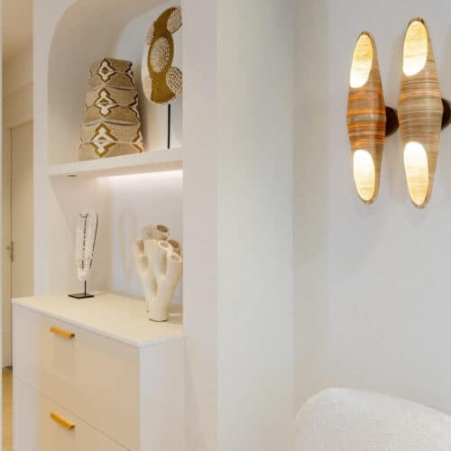 Impressie van een deel van een witte Japandi keuken. We zien een nis met ronde hoeken, met de uitstraling van een toog. De nis is voorzien van twee onderkasten met houten handgrepen, met daarboven een verlichte plank met decoraties. Naast de nis zien we twee organisch gevormde wandlampen.