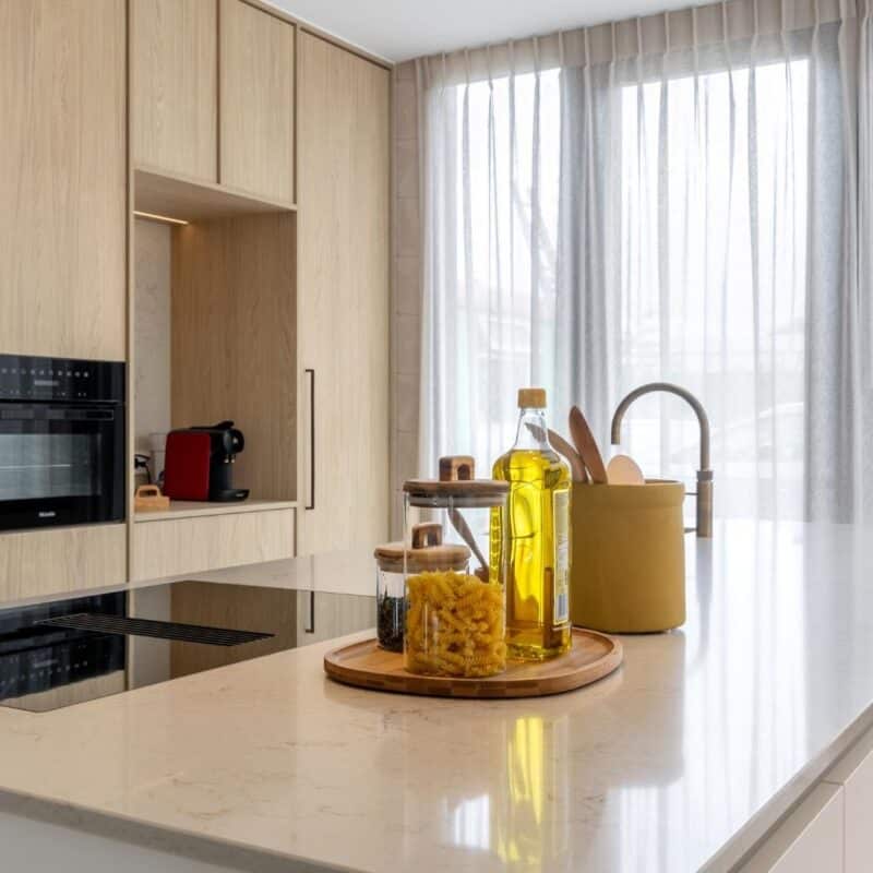 Impressie van een Japandi stijl keuken met kookeiland, natuursteenlook werkblad en licht eiken kastenwand met inbouw ovens.