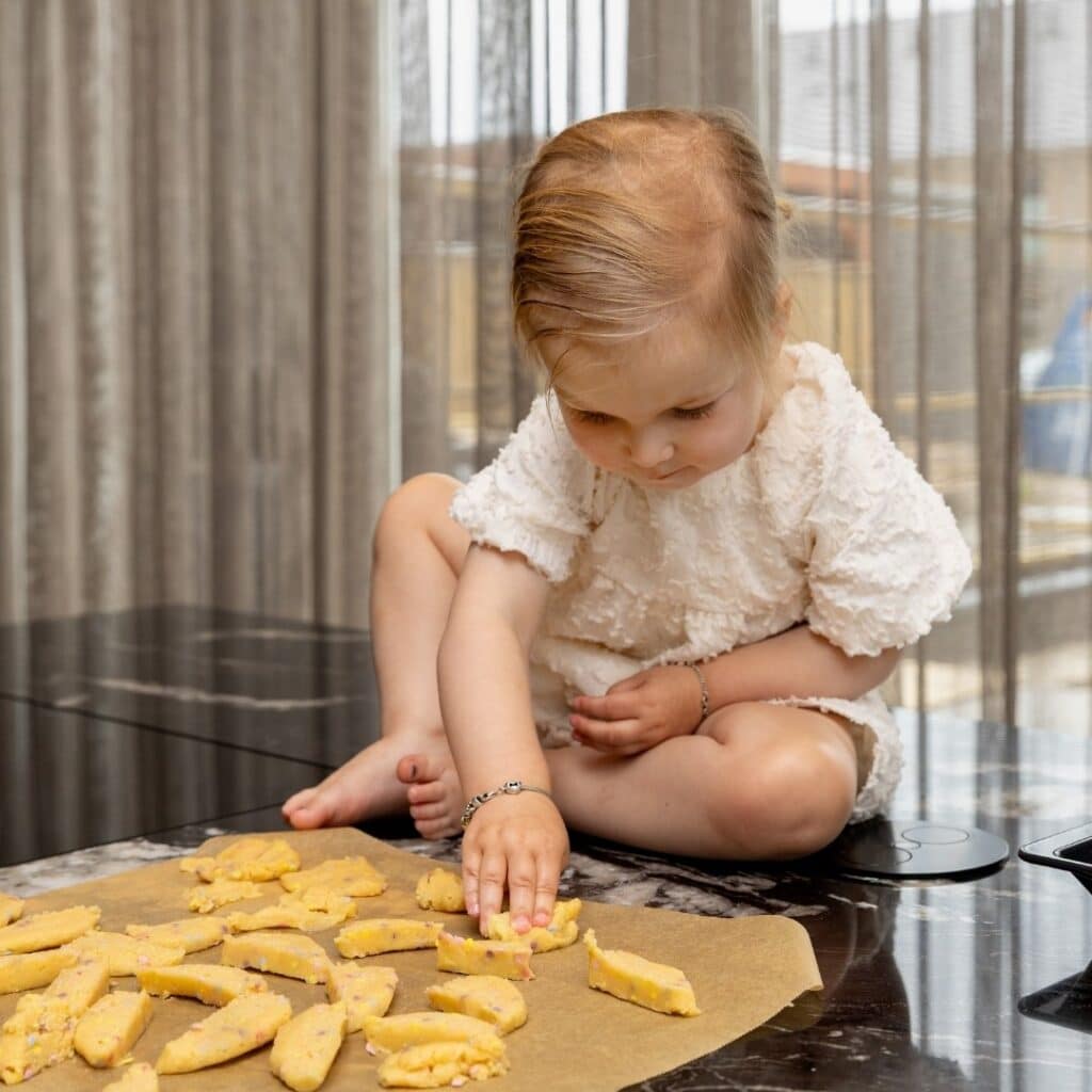 Impressie van een kind, zittend op een kookeiland met zwart marmerlook werkblad. Het kind heeft een bakvel met stukjes deeg voor zich liggen, en is een stukje aan het kneden.