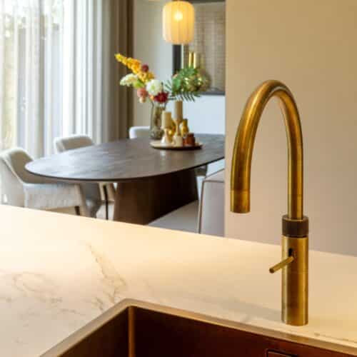 Impressie van een Quooker in messing patina finisch. Op de achtergrond zien we een eethoek met ovale houten tafel.
