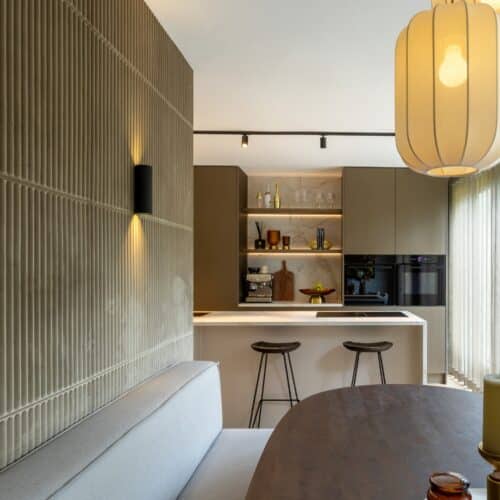 Kijk vanuit een eethoek naar een luxe keuken, voorzien van kookeiland met bar en een houten kastenwand met nis.