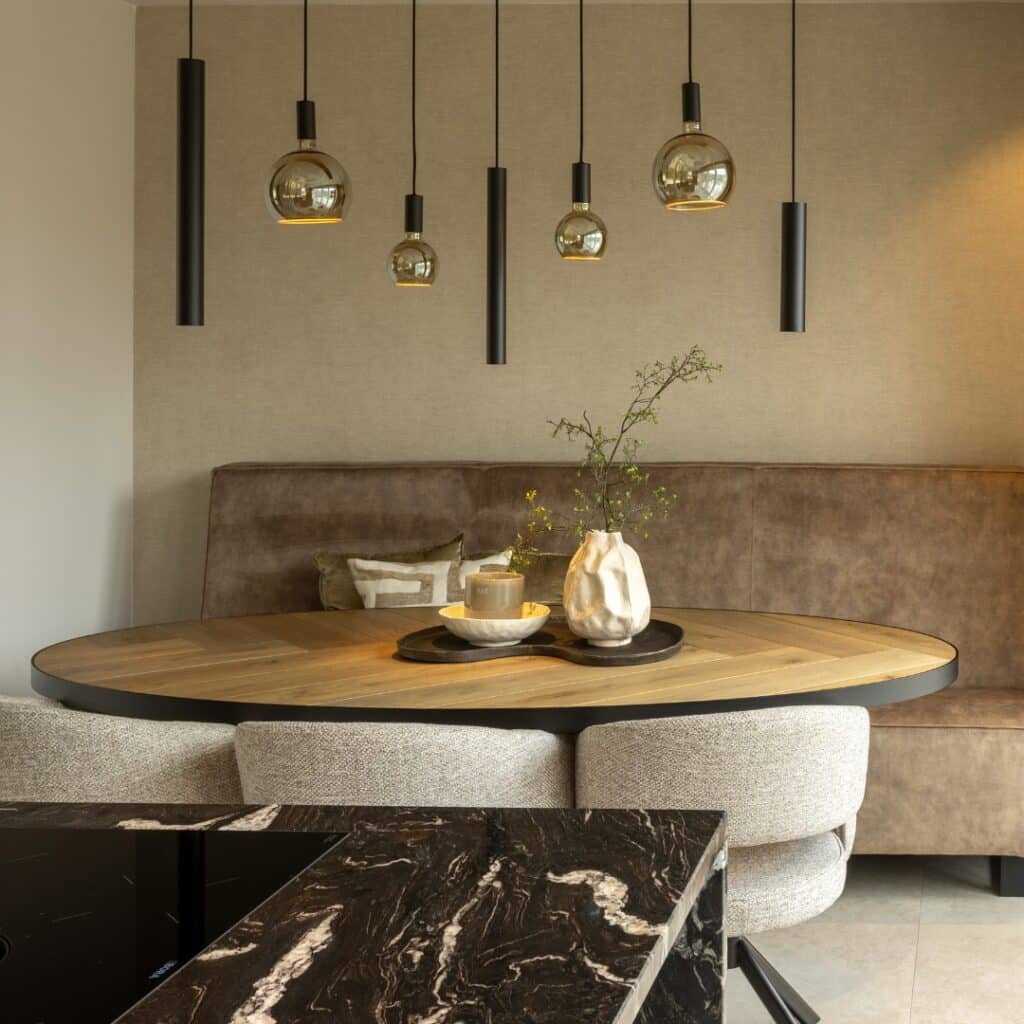 Hotel Chique leefruimte, voorzien van zithoek met ovale houten tafel, bank en drie stoelen. We kijken ernaar vanuit een moderne keuken met zwart marmerlook kookeiland.