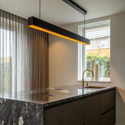 Impressie van een kookeiland met zwart marmerlook blad en zijwand, een Quooker met verweerde messing finish, een kookplaat met werkbladafzuiging en een zwarte designlamp boven het eiland met warm licht.