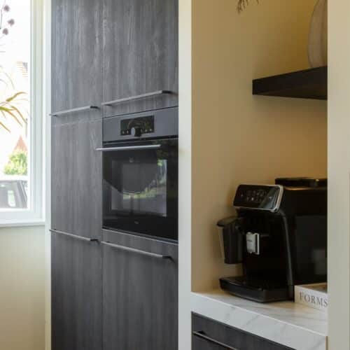 Impressie van een houten kastenwand in zwart, voorzien van een inbouwoven en nis met koffiemachine.