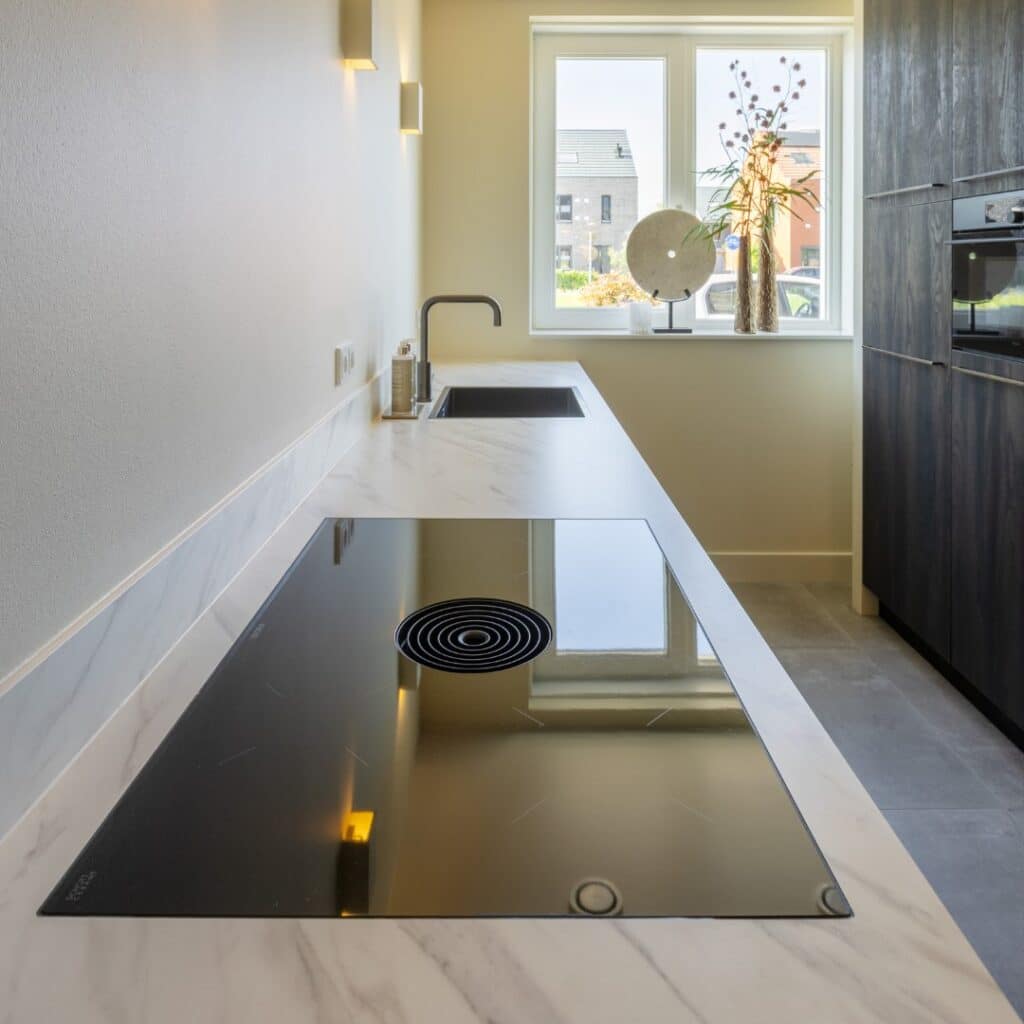 Impressie van een parallel keuken, met de focus op een natuursteenlook aanrecht met kookplaat met werkbladafzuiging en een Quooker.