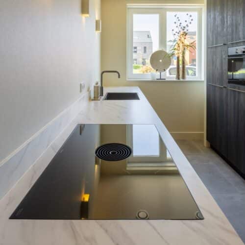 Impressie van een parallel keuken, met de focus op een natuursteenlook aanrecht met kookplaat met werkbladafzuiging en een Quooker.