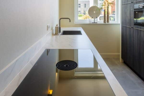 Impressie van een parallel keuken, met de focus op een natuursteenlook aanrecht met kookplaat met werkbladafzuiging en een Quooker.