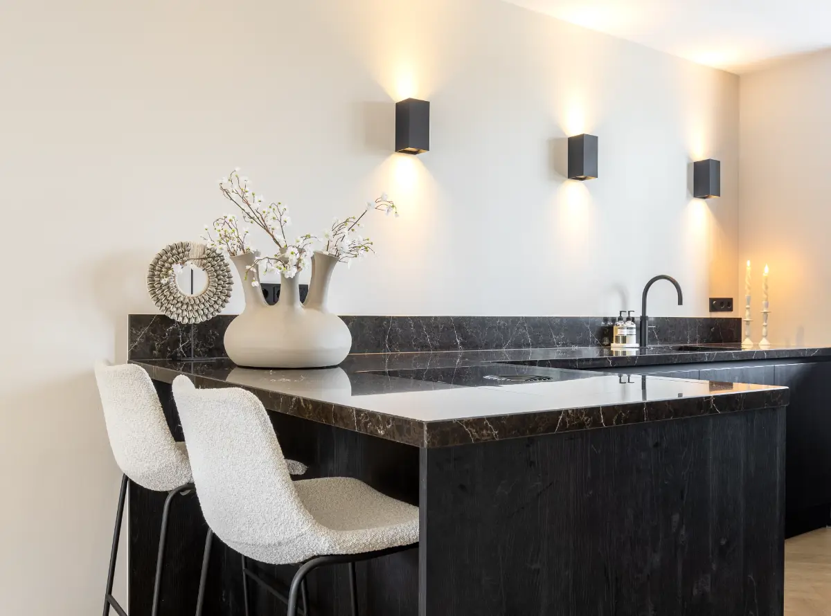 Zwart houten kookeiland met een donker marmeren blad, witte bouclé barkrukken en moderne zwarte wandlampen op een witte muur.