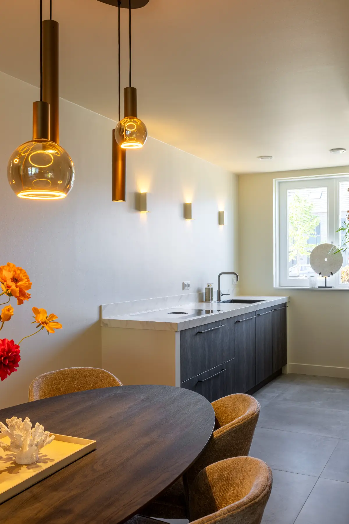 Modern keukenontwerp met donkere houten onderkasten, wit marmeren werkblad en luxe gouden hanglampen boven een houten tafel.