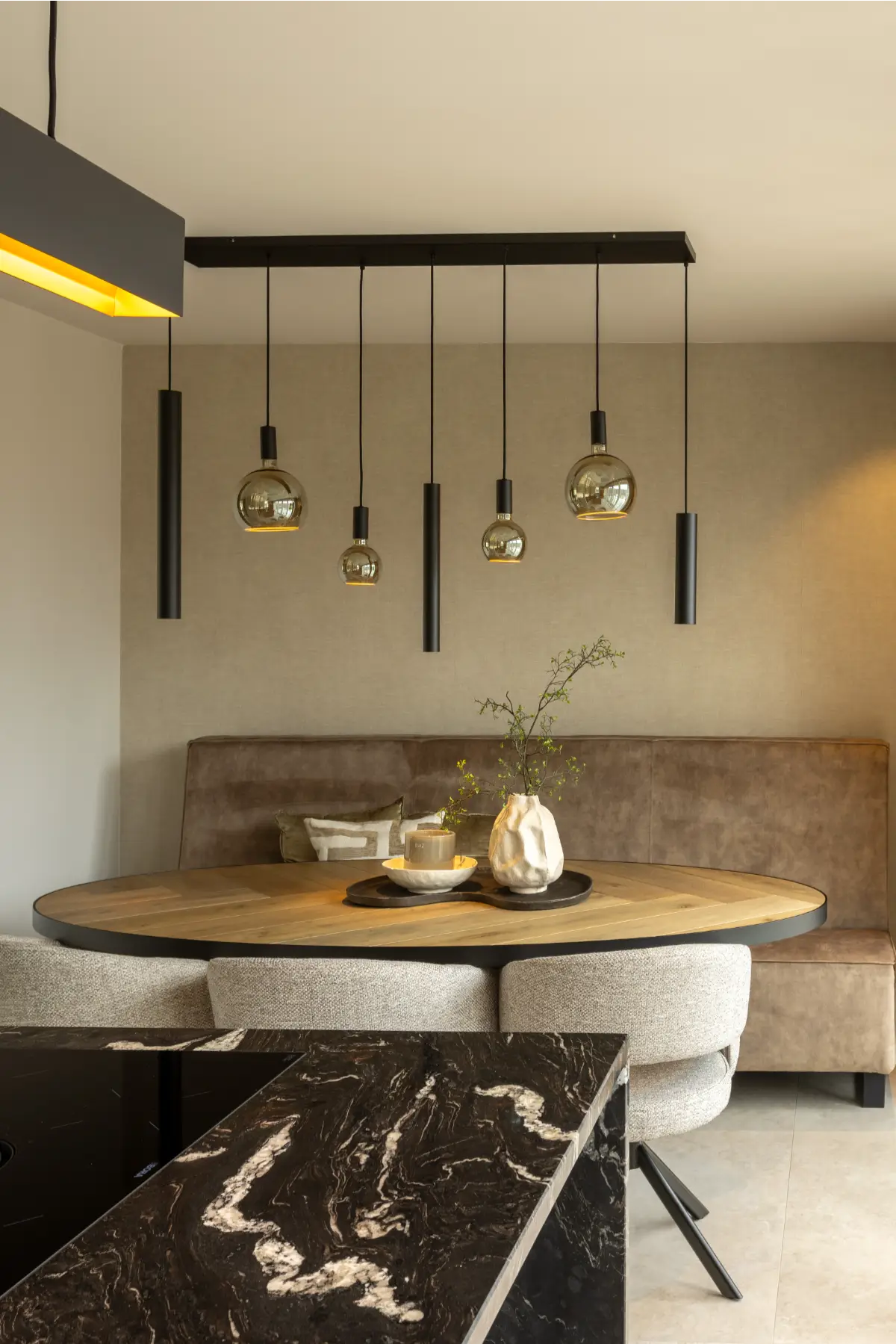 Detail van een ronde houten eettafel met een beige fluwelen zitbank en een unieke zwarte hanglamp met verschillende glazen bollen.