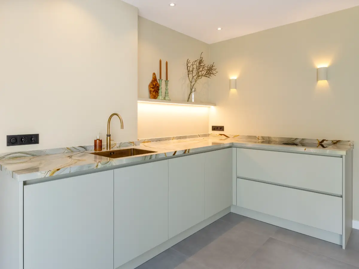 L-vormige lichtgroene keuken met een wit marmeren werkblad met bruine aders, een goudkleurige kraan en subtiele wandverlichting.